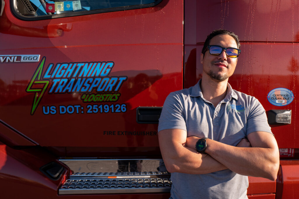 Man with glasses standing with arms crossed against drivers door of a red semi truck with lightning transport logo on the door